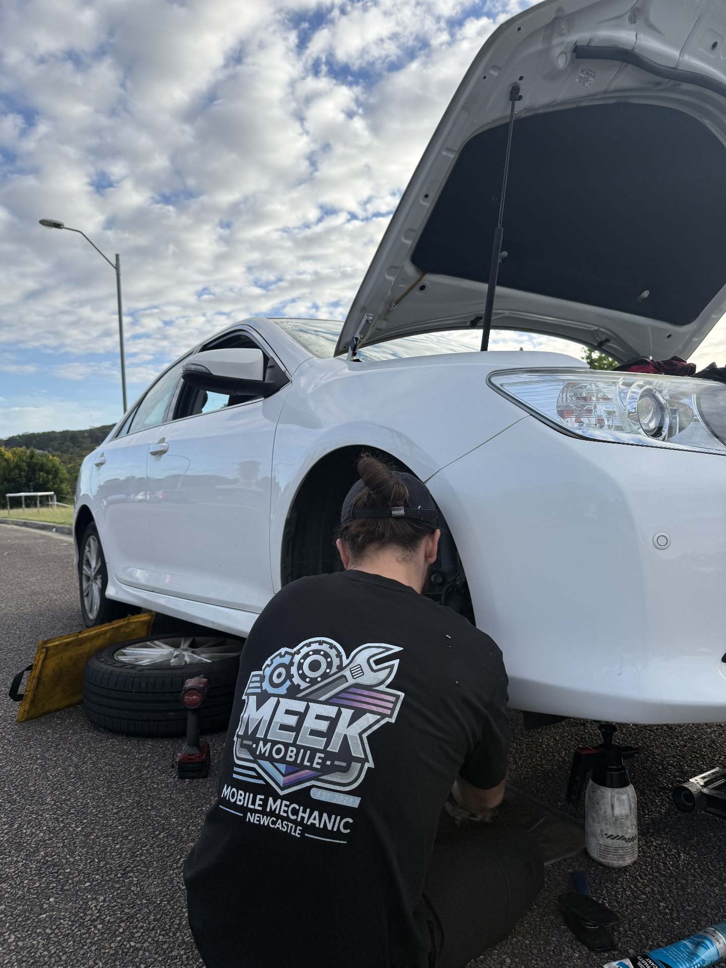 Meek Mobile mechanic kneeling beside a white sedan with the bonnet up, replacing a front wheel roadside in Newcastle
